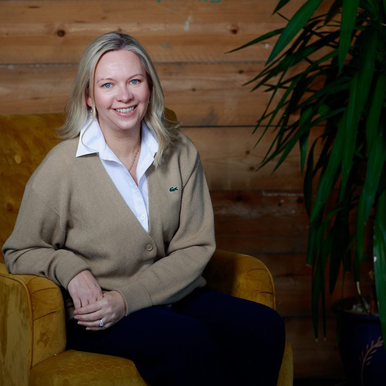 Smiling woman seated on a yellow armchair with a plant in the background.