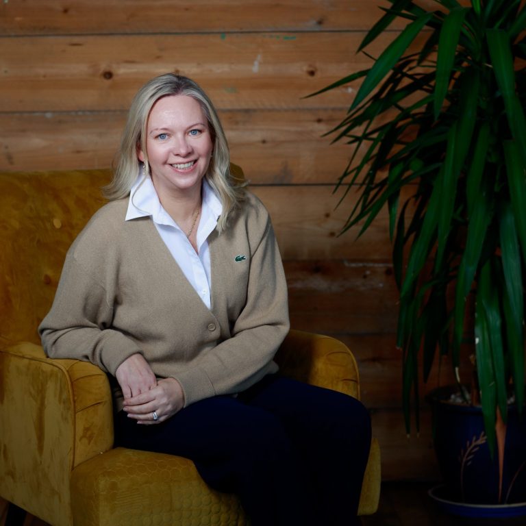 Woman holding a mug, sitting on a sofa with a laptop and plants in a sunny room.